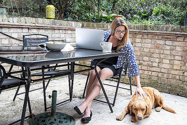 Schwangere Frau im mittleren Erwachsenenalter benutzt am Laptop auf dem Terrassentisch  während sie ihren Hund streichelt