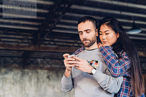 Frau schaut einem Mann beim Benutzen eines Smartphones über die Schulter