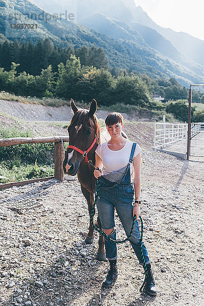 Junge Frau führt Pferd auf ländlichem Reitplatz  Ganzkörperporträt