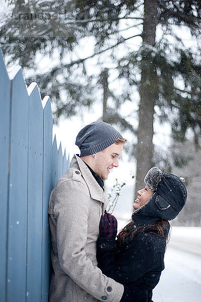 Junges Paar umarmt sich im Schnee gegen einen Zaun
