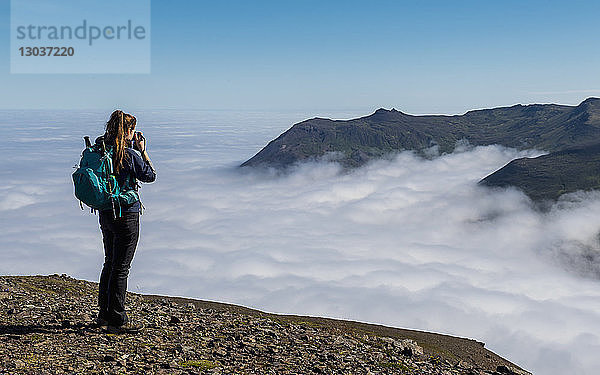 Ganzkörperaufnahme einer Wanderin  die den Blick auf die über dem Nebel aufragenden Berge fotografiert  Borgafjordur Eystri  Island