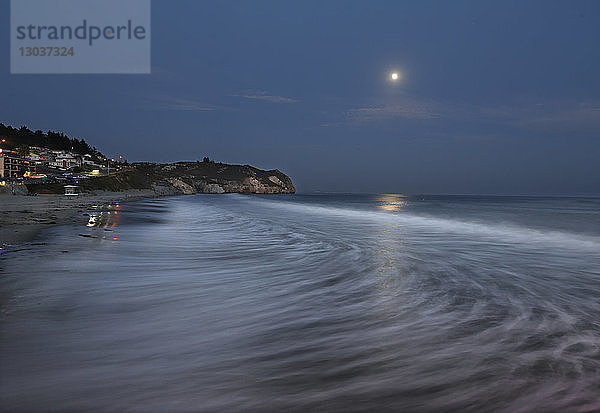 Eine Welle bricht sich im Pazifischen Ozean  während der Vollmond an einem Sommertag am Avila Beach in Kalifornien  USA  aufgeht.