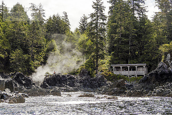 Ein Mann sitzt auf einem Felsen  während hinter ihm ein Strom aus einer heißen Quelle in Hot Springs Cove in Tofino  British Columbia  Kanada  entspringt. In der Nähe ist ein Umkleideraum eingerichtet  damit die Besucher ihre Badekleidung anziehen können  um in die heißen Quellen zu gehen.