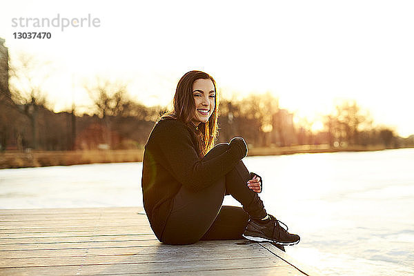 Seitenansicht einer einzelnen lächelnden jungen Frau  die bei Sonnenuntergang auf einem Steg sitzt