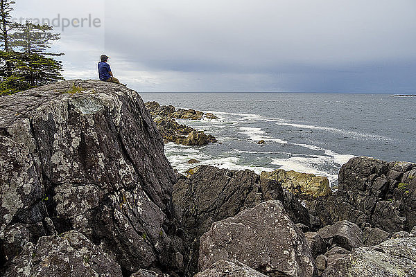 Blick auf einen einsamen Mann  der auf einer Felsformation am Meeresufer sitzt  Hot Springs Cove  Tofino  British Columbia  Kanada