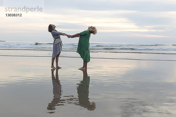 Glückliche Freundinnen in voller Länge halten sich bei Sonnenuntergang am Strand an den Händen  während sie am Ufer stehen