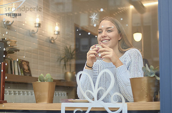Lächelnde Frau trinkt Kaffee  während sie zu Weihnachten durch ein Fenster gesehen am Tisch im Café sitzt