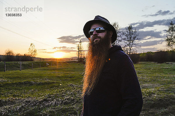 Bärtiger Mann mit Hut und Sonnenbrille schaut weg  während er bei Sonnenuntergang auf dem Feld gegen den Himmel steht