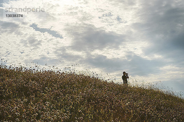 Wanderer schaut durch ein Fernglas  während er auf einem Hügel vor bewölktem Himmel steht
