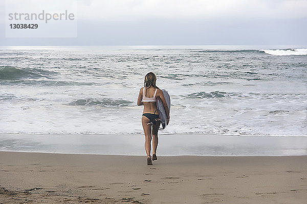 Rückansicht einer Frau  die ein Surfbrett trägt  während sie am Strand auf das Meer zuläuft