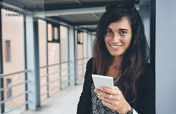 Porträt einer lächelnden Frau  die ein Mobiltelefon benutzt  während sie auf einer Fußgängerbrücke steht
