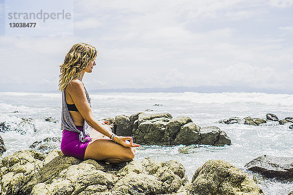 Seitenansicht einer mittleren erwachsenen Frau  die in Lotus-Position auf Felsen am Strand meditiert