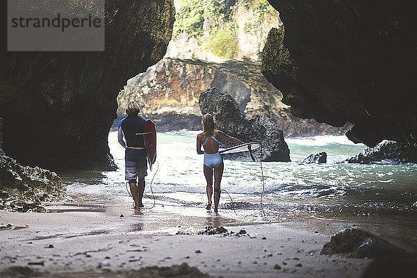 Rückansicht von Freunden  die beim Strandspaziergang an Felsformationen ein Surfbrett tragen