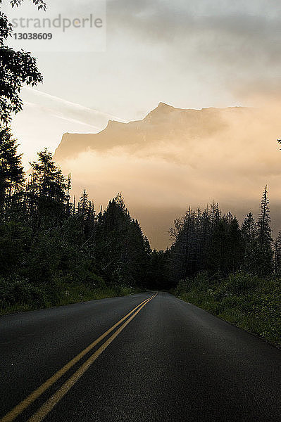 Leere Straße inmitten von Bäumen gegen bewölkten Himmel im Glacier National Park bei Sonnenuntergang