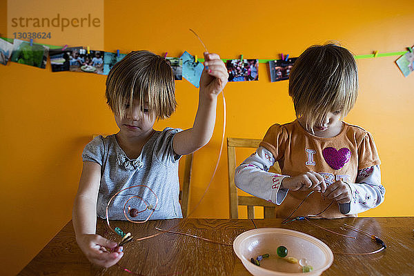 Schwestern machen Perlendekoration bei Tisch  während sie zu Hause an der Wand stehen