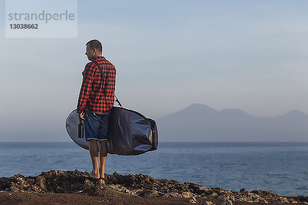 Seitenansicht eines Mannes  der eine Tasche trägt  während er bei Sonnenuntergang am Strand gegen den Himmel steht