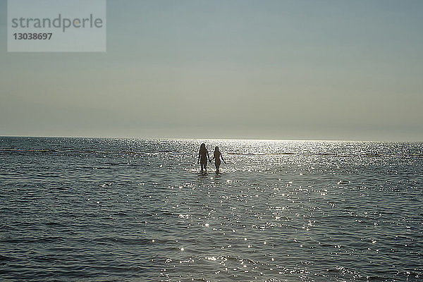 Mutter und Tochter gehen als Silhouette im Meer  während sie die Hände gegen den Himmel halten