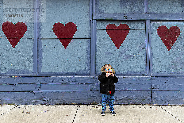 Junge in voller Länge isst Nahrung  während er auf dem Fußweg gegen eine am Herzen gezogene Wand steht
