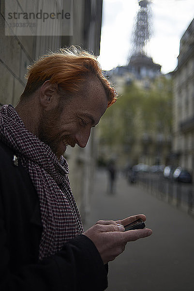 Seitenansicht eines lächelnden männlichen Touristen mit einem Smartphone auf der Straße mit dem Eiffelturm im Hintergrund