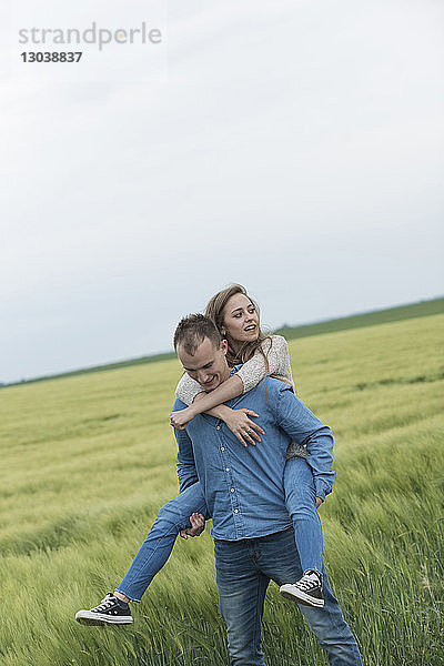 Junger Mann nimmt Frau huckepack auf Grasfeld gegen den Himmel