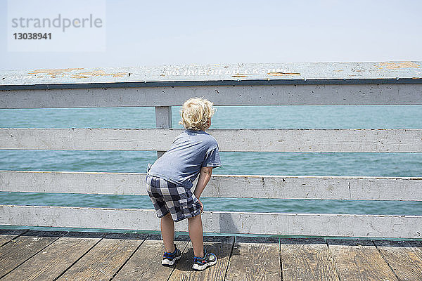 Rückansicht eines Jungen  der durch den Zaun schaut  während er sich am Pier beugt