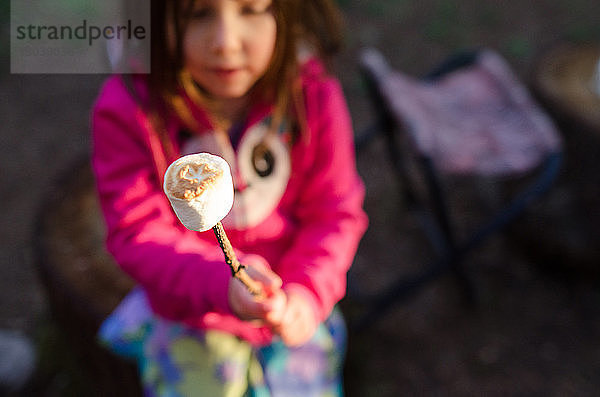 Schrägaufnahme eines Mädchens  das Marshmallows am Stock hält  während es auf einem Campingplatz sitzt