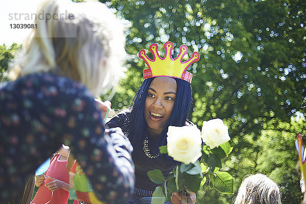 Glückliche Frau trägt Papierkrone  während sie während der Geburtstagsfeier im Hinterhof einem Teenager-Mädchen weiße Rosen schenkt