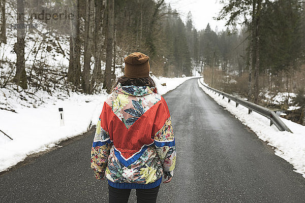 Rückansicht einer Frau in warmer Kleidung  die im Winter auf einer Landstraße zwischen Bäumen im Wald steht