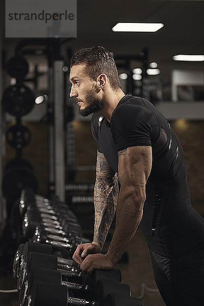Seitenansicht eines selbstbewussten Mannes  der sich beim Training im Fitnessstudio auf Hanteln stützt