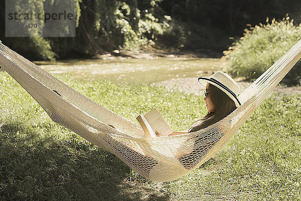 Frau liest Buch beim Entspannen auf Hängematte im Park
