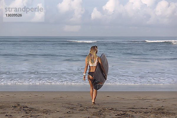 Frau in voller Länge  die ein Surfbrett trägt  während sie am Strand in Richtung Meer läuft