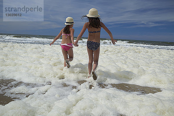 Rückansicht von Schwestern  die in Meeresschäumen laufen  während sie am Strand Händchen halten