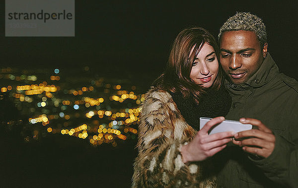 Glückliches junges Paar betrachtet Smartphone  während es sich gegen das Stadtbild stellt
