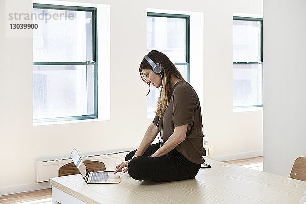Seitenansicht einer Geschäftsfrau  die in einem kreativen Büro am Schreibtisch sitzend über einen Laptop Musik hört