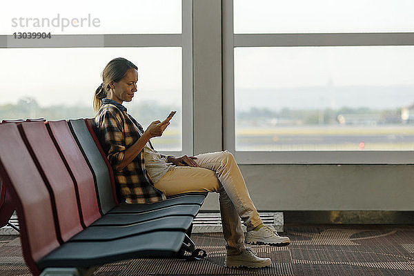 Seitenansicht einer lächelnden Frau beim Benutzen eines Mobiltelefons im Abflugbereich des Flughafens