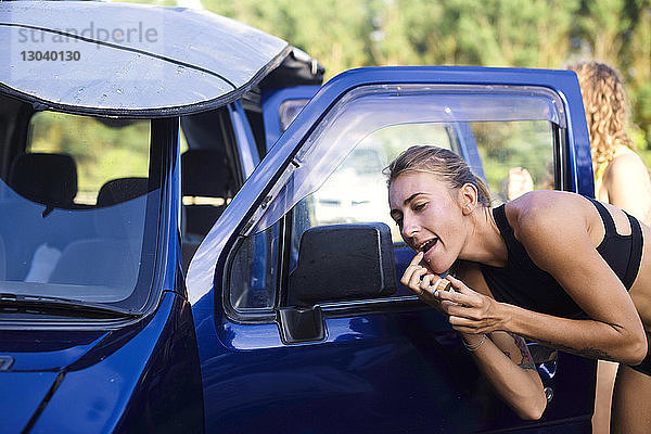 Frau trägt Lippenbalsam auf  während sie in den Seitenspiegel eines Autos schaut