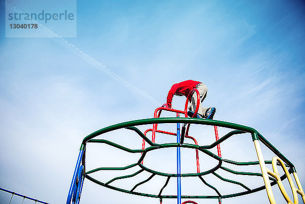 Niedrigwinkelansicht eines Jungen  der auf Affenstangen gegen den Himmel auf einem Spielplatz klettert