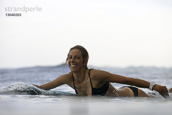 Porträt einer glücklichen Frau  die auf einem Surfbrett am Strand gegen den Himmel liegt