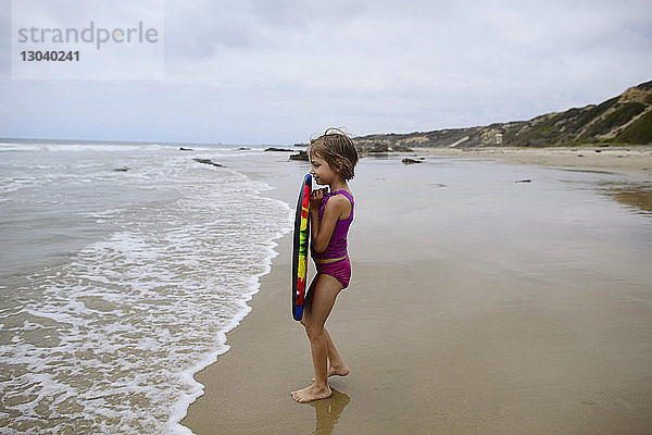 Seitenansicht eines Mädchens  das ein Surfbrett hält  während es am Strand des Crystal Cove State Park gegen den Himmel steht