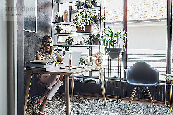 Glückliche Geschäftsfrau  die am Laptop arbeitet  während sie im Heimbüro am Tisch sitzt