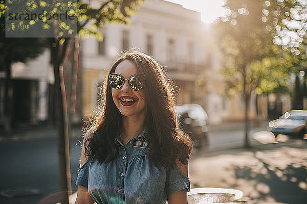 Fröhliche junge Frau mit Sonnenbrille  die am sonnigen Tag auf dem Bürgersteig in der Stadt steht