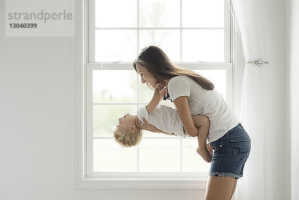 Seitenansicht einer Mutter  die einen Sohn trägt  während sie zu Hause gegen ein Fenster spielt