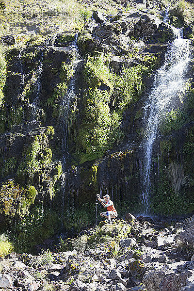 Wanderer  der bei Sonnenschein auf Felsen gegen Wasserfall und Berg kauert