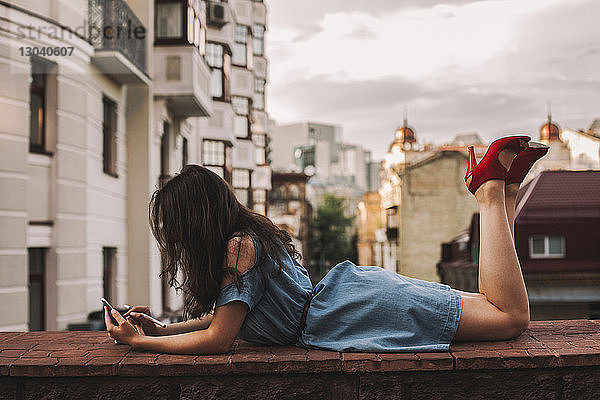 Seitenansicht einer jungen Frau mit erhobenen Füßen  die ein Smartphone benutzt  während sie auf einer Stützmauer gegen Gebäude in der Stadt liegt