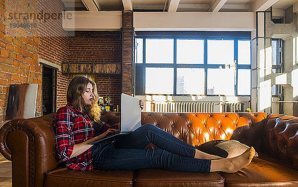 Junge Frau in voller Länge mit Laptop-Computer  während sie zu Hause auf einem Ledersofa sitzt
