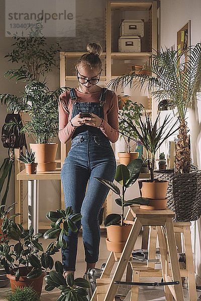 Frau benutzt Mobiltelefon  während sie sich zu Hause an Topfpflanzen an den Tisch lehnt