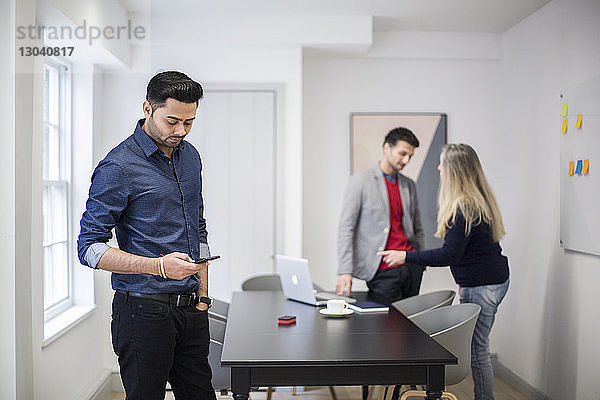 Geschäftsmann nutzt Smartphone mit Kollegen  die im Hintergrund am Schreibtisch stehen