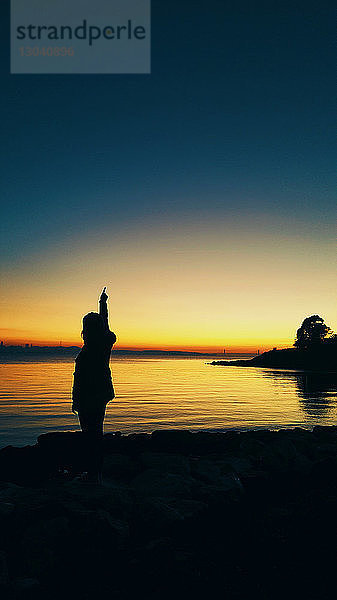 Silhouette eines Teenager-Mädchens mit erhobener Hand  das bei Sonnenuntergang am Strand vor klarem Himmel steht