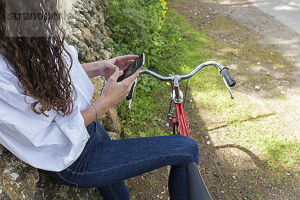 Mittelsektion einer Frau  die ein Mobiltelefon benutzt  während sie mit dem Fahrrad an einer Steinmauer steht