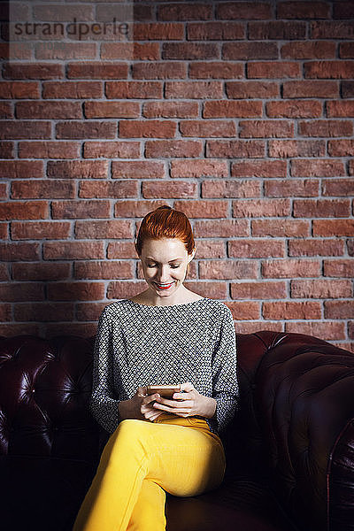Glückliche Geschäftsfrau benutzt Smartphone  während sie in der Büro-Lobby auf der Couch sitzt